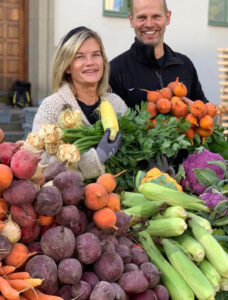 Leende kvinna och man och ett marknadsstånd fylld av grönsaker i olika färger.
