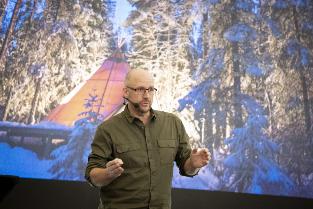 En man håller föredrag i bakgrunden är bild på upplyst kåta i snölandskap.